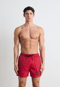 Un short de bain rouge en matériau léger avec une taille élastique et un cordon noir. Logo "HUGO" imprimé en noir sur le côté.