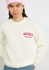 Pull en tricot ivoire clair avec un col ras du cou côtelé, arborant une broderie rouge "DICKIES" et "Quality Made" sur la poitrine gauche. Texture douce.