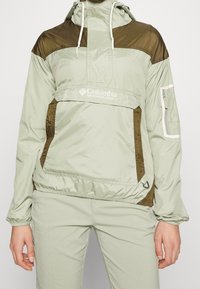 Persona con chaqueta cortavientos de color verde claro y marrón de Columbia con capucha y pantalones a juego, de pie contra un fondo simple.