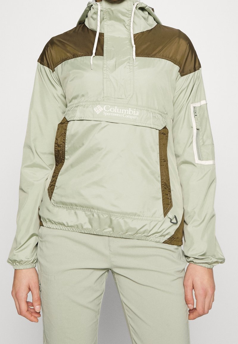 Persona con chaqueta cortavientos de color verde claro y marrón de Columbia con capucha y pantalones a juego, de pie contra un fondo simple.