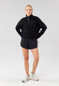 Černý fleecový pulovr s zipem na krku, v kombinaci s černými šortkami a bílými teniskami. Pulovr má volný střih a žebrovaný lem.