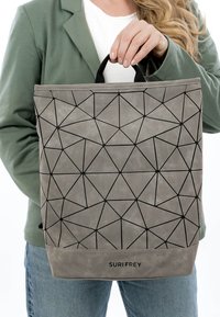 Mujer con chaqueta verde sosteniendo un bolso Suri Frey de patrón geométrico gris por su asa negra.