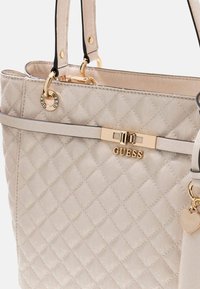 Borsa tote trapuntata beige con hardware dorato e logo "GUESS". Presenta doppi manici, dettagli decorativi e una texture liscia.
