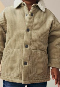 Veste en velours côtelé beige avec un col en shearling, des boutons-pression noirs, deux poches avant et une texture côtelée.