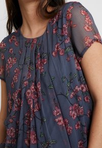 Blommig blus i genomskinligt marinblått tyg, med rosa och gröna blommor, korta ärmar, veckad design och rund halsringning.