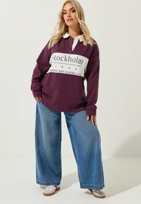 Lila långärmad polo med vit krage. Har en framsida med grafiken "Stockholm 1998 Sports and Leisure Club." Matchad med vida denimbyxor.