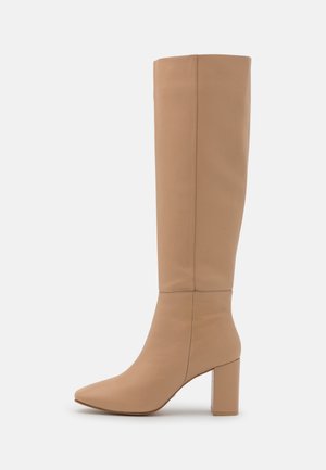 Botte en cuir marron clair montant jusqu'au genou, avec talon bloc, bout pointu et détail de couture verticale et autour de la cheville, présentée sur fond blanc.
