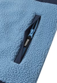 Tissu en polaire bleu clair avec une surface texturée, comportant une poche zippée bleu marine avec un tirette bleu et une étiquette de marque.