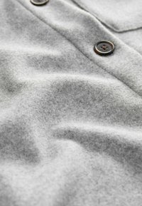 Grauer Stoff mit weicher Textur und leichtem Glanz, versehen mit dunklen Knöpfen und dezenten Falten, die auf ein Kleidungsstück hinweisen.