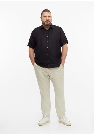 Man with beard and tied-back hair stands with hands in pockets wearing black short-sleeve button shirt, beige pants, and white sneakers.