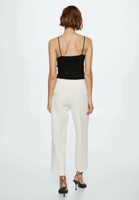 Femme aux cheveux bruns courts, portant un haut à bretelles spaghetti noir, un pantalon blanc taille haute et des sandales à talons noires, de dos.