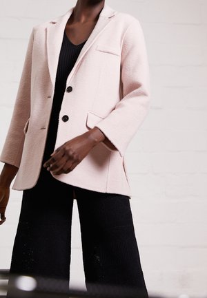 Vrouw draagt een lichtroze getextureerde blazer met zwarte knopen over een zwart geribd topje en wijde zwarte broek met gestructureerd patroon.