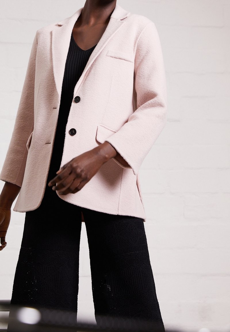 Femme portant un blazer texturé rose pâle avec des boutons noirs, un top noir côtelé et un pantalon noir large à motif texturé.