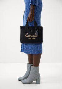 Black fabric tote bag with "Coach" and "New York" embroidered in gold, featuring double handles and a hang tag, paired with light grey ankle boots.