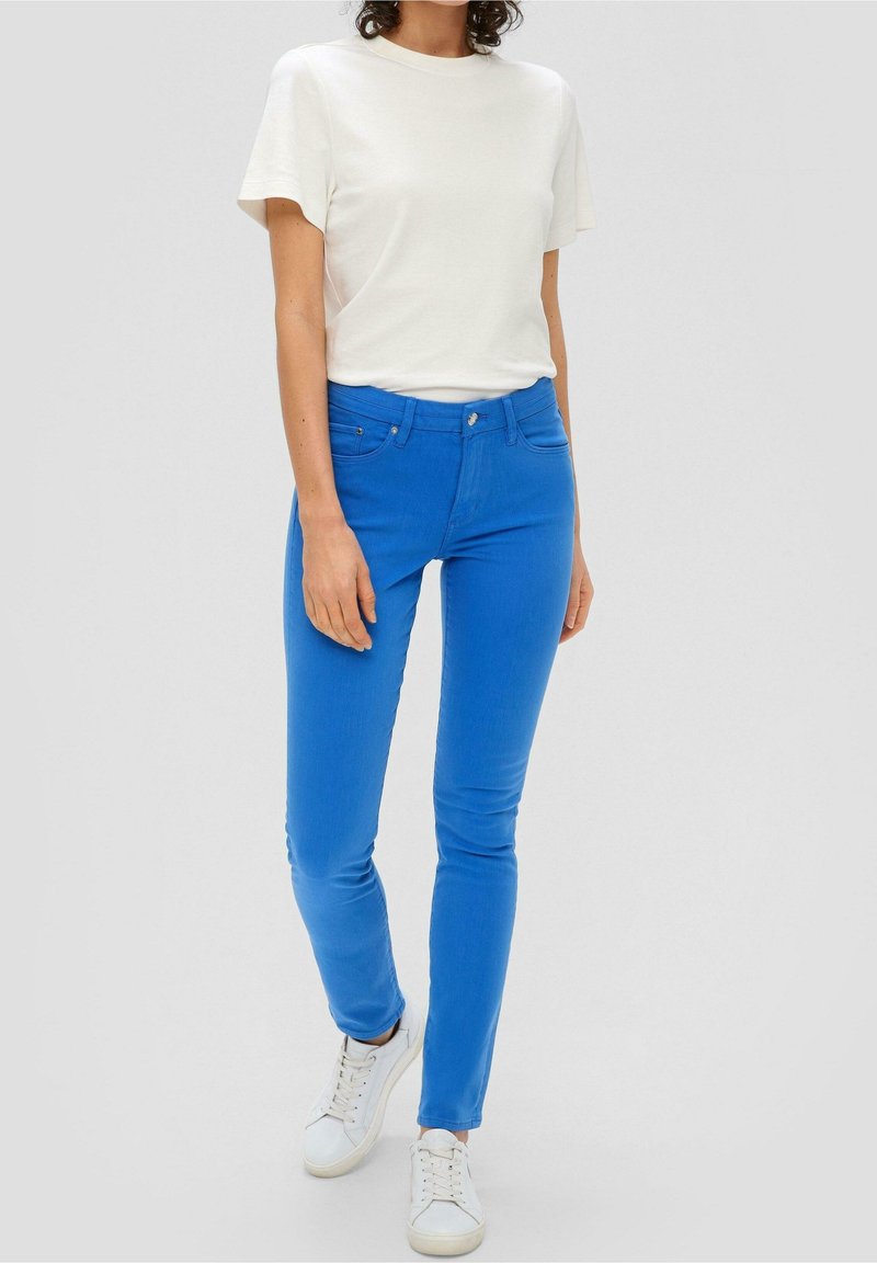 Personne portant un t-shirt blanc uni à manches courtes, un pantalon slim bleu vif et des baskets blanches, debout sur un fond gris clair.