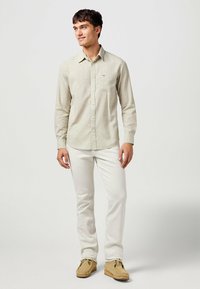 Camicia beige a maniche lunghe con bottoni e taschino sul petto, abbinata a pantaloni bianchi e scarpe mocassino color cammello. Tessuto morbido, vestibilità casual.