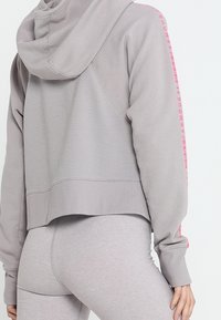 Persona che indossa un hoodie corto grigio chiaro con schiena a coste e leggings coordinati, la manica dell'hoodie mostra il logo rosa di Under Armour.