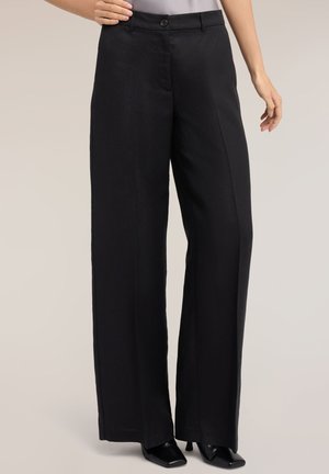 Femme portant un pantalon noir à jambes larges avec pli devant, fermeture par bouton et zip, associé à des talons carrés noirs.