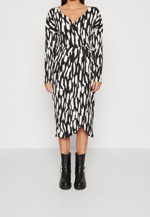 Kobieta ubrana w czarno-białą, abstrakcyjną, wzorzystą sukienkę midi z długim rękawem i dekoltem w serek oraz czarne botki z klamrami.