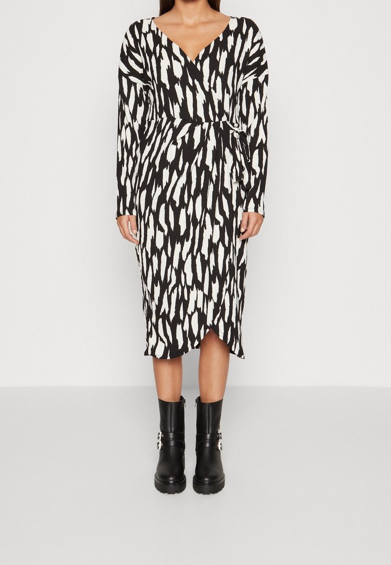 Femme portant une robe midi à manches longues avec un motif abstrait noir et blanc, un col en V, et des bottines noires à boucles.