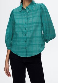 Blusa transparente de cuadros color verde azulado con botones y mangas abullonadas de tres cuartos, combinada con pantalones negros, recortada a la altura del cuello y a mitad del muslo.