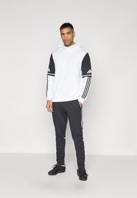 Kapuzensweatshirt in Weiß mit schwarzen Ärmeln, ausgestattet mit drei Streifen an jedem Arm; kombiniert mit dunkelgrauen Hosen und schwarzem Schuhwerk.