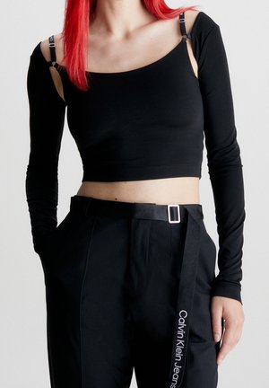 Personne aux cheveux roux portant un crop top noir épaules dénudées, un pantalon noir et une ceinture Calvin Klein Jeans sur un fond clair.