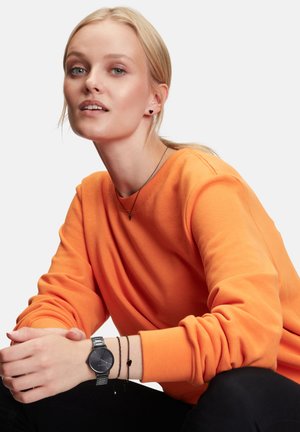 Orangener Sweatshirt, silberne Uhr mit schwarzem Ziffernblatt, kombiniert mit einem dunklen Armband. Glatter Stoff, lässige Passform, schlichtes Design, minimale Hardware.