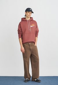 Μπορντό φούτερ με λευκό λογότυπο, μπροστινή τσέπη· καφέ παντελόνια με ευρύ μπατζάκι, σκούρα αθλητικά παπούτσια· το μοντέλο φοράει μπλε σκούφο baseball. Απλό φόντο.