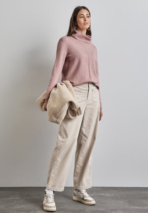 Hellrosa Rollkragenpullover, beige Sherpa-Jacke, cremefarbene, weite Hose und beige-weiße Sneakers vor einem neutralen Hintergrund.