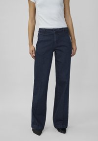 Een vrouw staat in een aansluitend wit T-shirt en een donkerblauwe wijde jeans, gecombineerd met zwarte puntige hakken, voor een neutrale achtergrond.