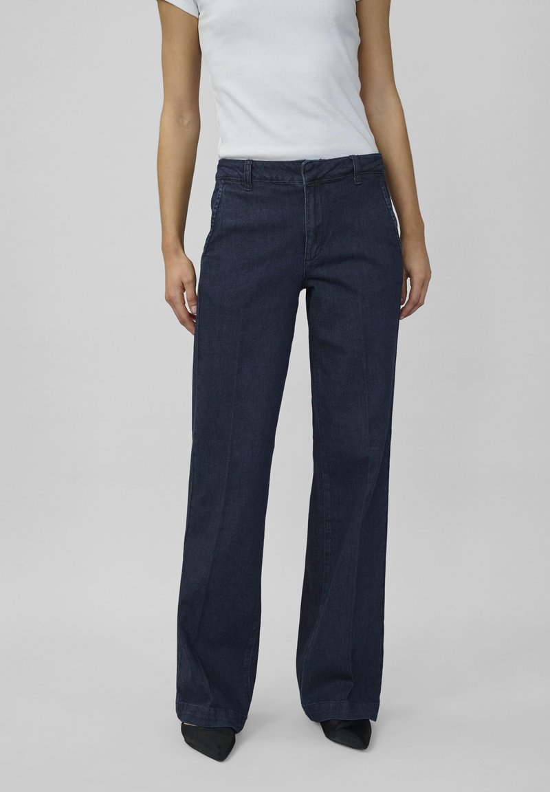 Een vrouw staat in een aansluitend wit T-shirt en een donkerblauwe wijde jeans, gecombineerd met zwarte puntige hakken, voor een neutrale achtergrond.