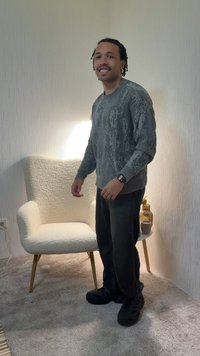 Νεαρός άνδρας που χαμογελά, φορώντας γκρι πουλόβερ και μαύρο παντελόνι, στέκεται δίπλα σε μια λευκή πολυθρόνα και ένα μικρό στρογγυλό τραπεζάκι με διακοσμητικά.