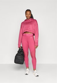 Różowy, krótki hoodie i dopasowane legginsy z teksturowanymi akcentami. Czarna torba sportowa, białe sneakersy. Gładkie i dopasowane materiały przez całość.