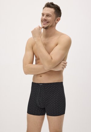 Hombre sonriente sin camiseta, de pie con los brazos cruzados, vistiendo calzoncillos bóxer negros con pequeños patrones blancos sobre un fondo claro y liso.