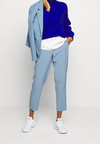 Person i blå skräddarsydda byxor, blå stickad tröja, vit skjorta, vita sneakers, som håller en matchande blå kavaj över axeln.