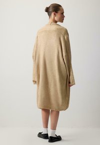 Beżowy, oversize'owy sweter dzianinowy z opadającymi ramionami, długimi rękawami, ściągaczami na mankietach i luźnym krojem, w połączeniu z czarnymi butami i białymi skarpetkami.