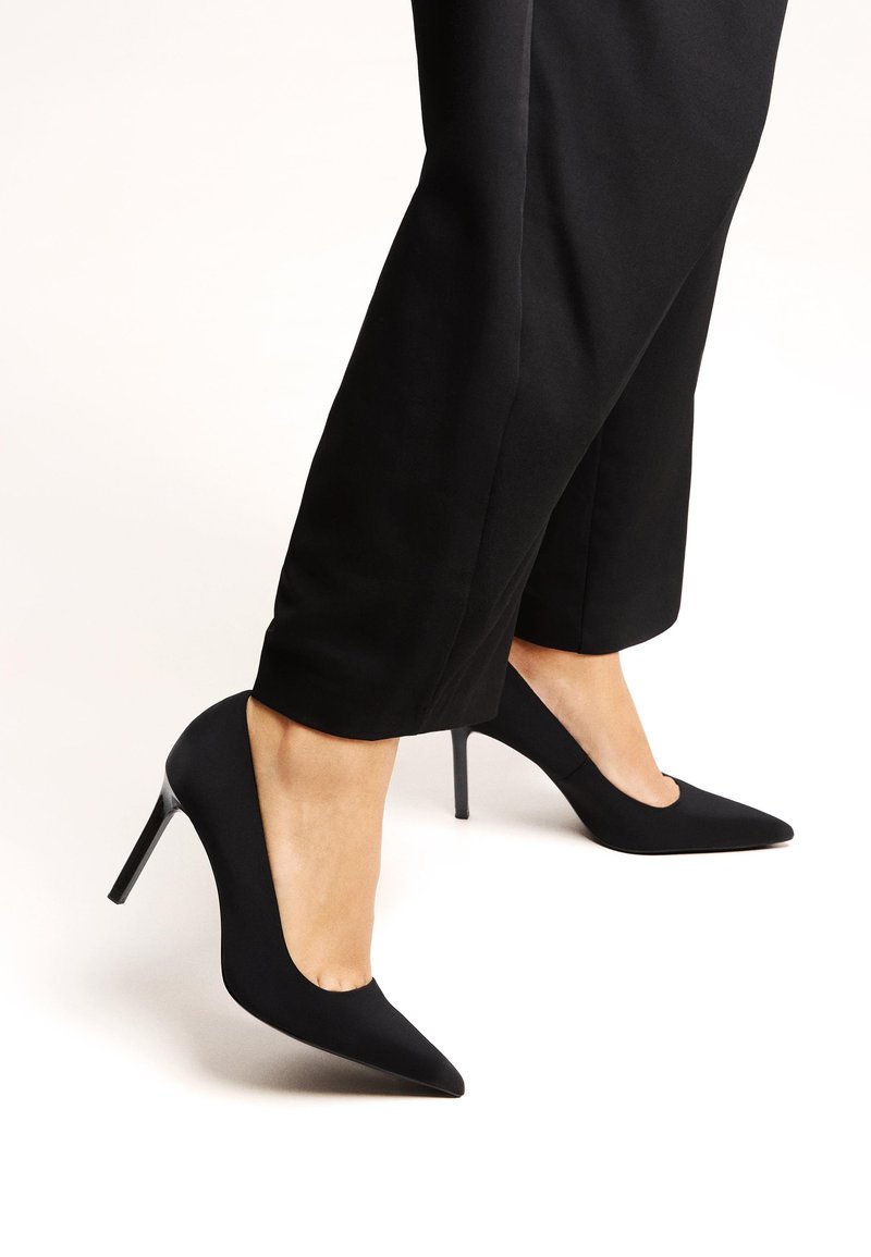 Black pointed-toe high heels with a matte finish, featuring a slim stiletto heel and a smooth texture, paired with cropped black trousers.