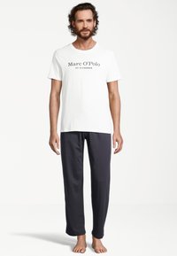 Weißes Baumwoll-T-Shirt mit schwarzem Aufdruck, auf dem "Marc O'Polo EST. IN STOCKHOLM" steht, kombiniert mit anthrazitfarbenen Jogginghosen. Barfuß.