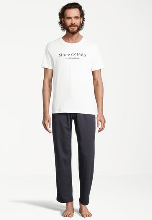 Weißes Baumwoll-T-Shirt mit schwarzem Aufdruck, auf dem "Marc O'Polo EST. IN STOCKHOLM" steht, kombiniert mit anthrazitfarbenen Jogginghosen. Barfuß.