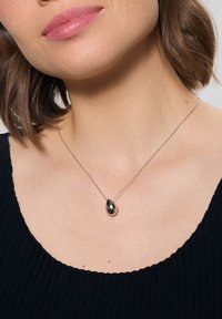 Kobieta z włosami do ramion ma na sobie czarną bluzkę oraz srebrny naszyjnik z zawieszką w kształcie kropli. Zbliżenie na jej dekolt.