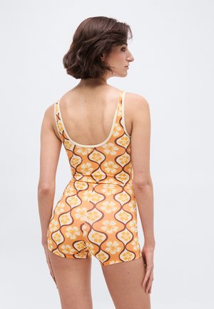 Γυναίκα με κοντά καστανά μαλλιά που φοράει πορτοκαλί ολόσωμη φόρμα με λουλουδάτο μοτίβο χωρίς μανίκια, όρθια με την πλάτη προς την κάμερα.