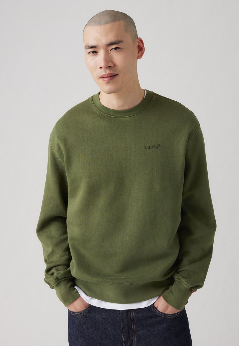 Sweatshirt vert à col rond en matériau doux, doté de poignets et d'un ourlet côtelés, avec un petit logo sur la poitrine. Porté sur une chemise blanche.