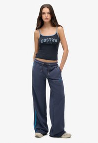 Jongedame met lang bruin haar, gekleed in een marineblauwe crop tanktop met "Boston" en losse donkerblauwe sweatpants met sneakers.