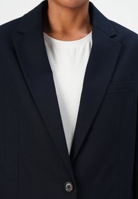Personne portant un blazer bleu marine foncé avec un seul bouton par-dessus un t-shirt blanc uni à col rond, visible du cou au milieu du torse.