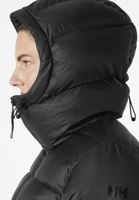 Profil d'une personne portant une veste matelassée noire avec une grande capuche zippée, montrant le logo Helly Hansen sur l'épaule.