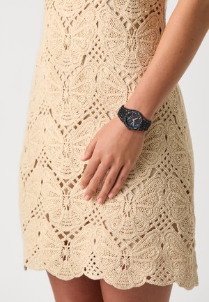 Montre colorée avec un bracelet noir, dotée d'un cadran multicolore. Portée sur une main près d'une robe en crochet beige avec des motifs de papillons.