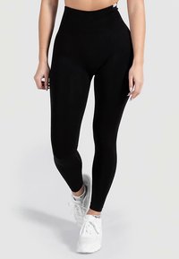 Zwarte leggings met hoge taille gemaakt van glad, rekbaar materiaal, slim fit ontwerp en subtiele branding bij de taille, gecombineerd met witte sneakers.