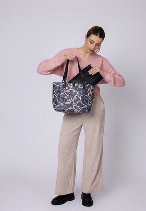 Borsa a spalla blu navy con motivo astratto dorato, dettagli neri e doppi manici, aperta da una persona che indossa un maglione rosa e pantaloni beige.