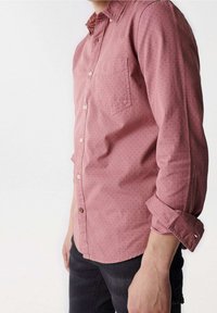Camicia button-up color rosa con taschino sul petto, texture a motivo sottile, maniche arrotolate e bottoni marroni. Indossata con jeans scuri.
